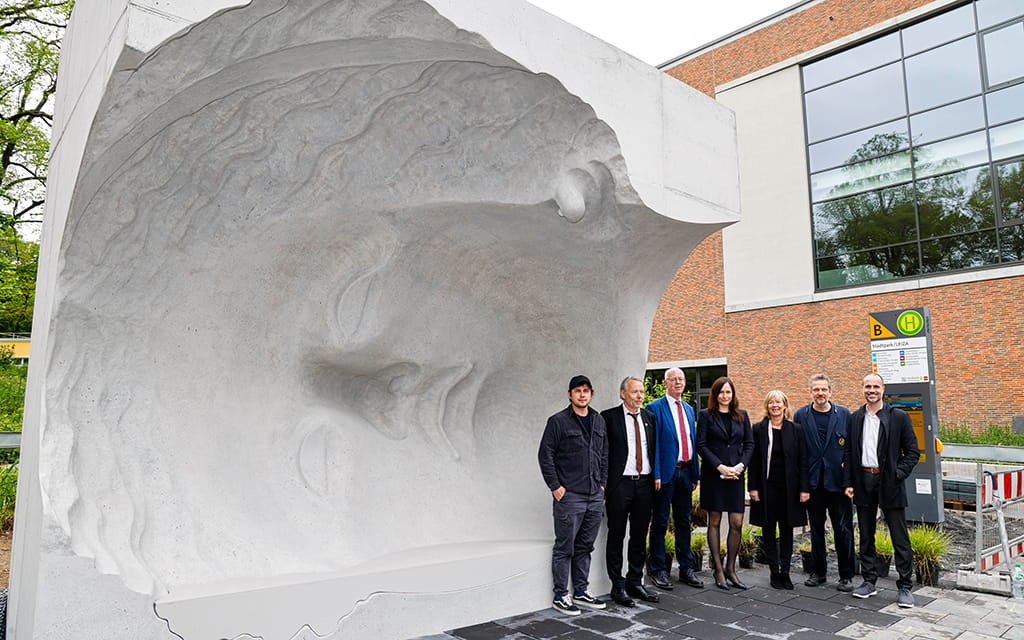Personengruppe vor Bushaltestelle aus Beton in Form eines großen Kopfes