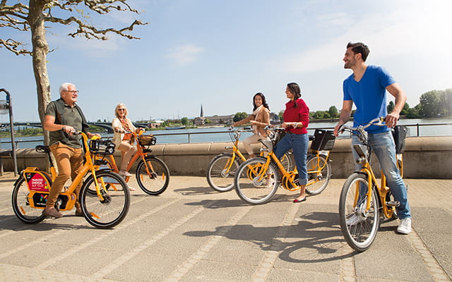 Gruppe Menschen steht mit Mieträdern der Mainzer Mobilität am Rhein 2 Fünf Menschen Treffen sich mit Fahrrädern von meinRad am Rheinufer