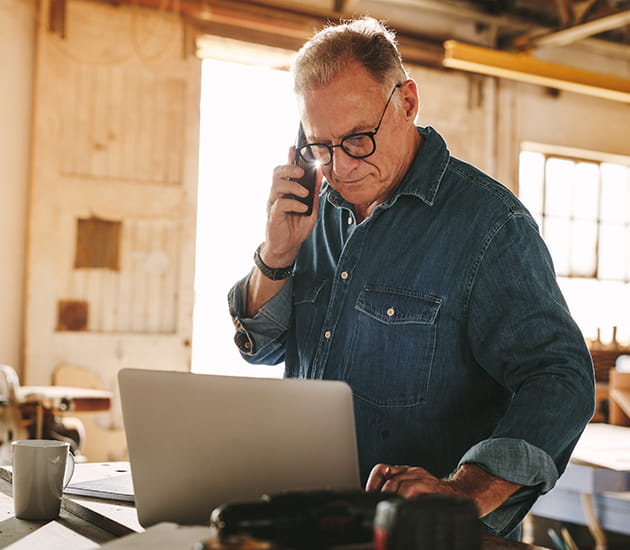 Älterer Herr steht telefonierend in einer Werkstatt und arbeitet an einem Laptop