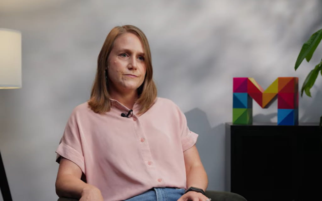 Blonde Frau mit rosa Bluse sitzt auf Stuhl mit buntem M im Hintergrund