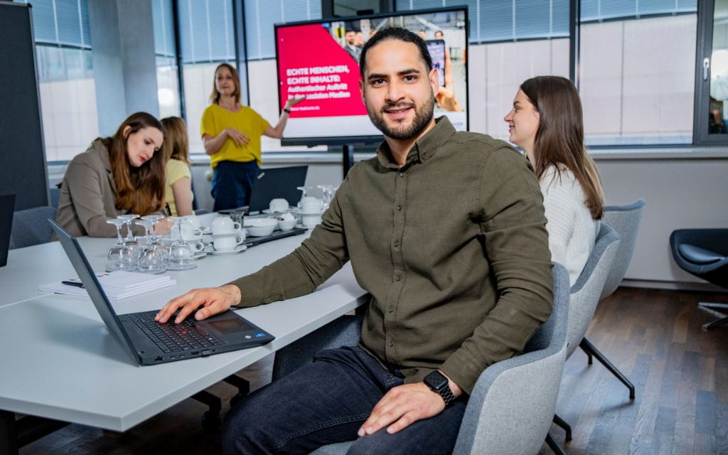 Ein Mann am Meetingtisch mit Laptop blickt in Kamera